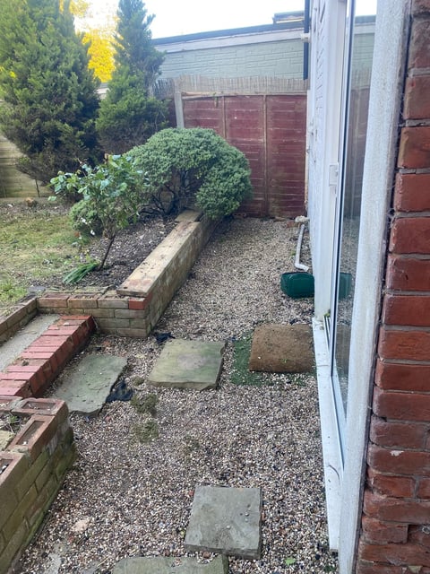 Narrow backyard garden with small potted shrub, gravel ground, concrete pavers, brick walls, and fencing