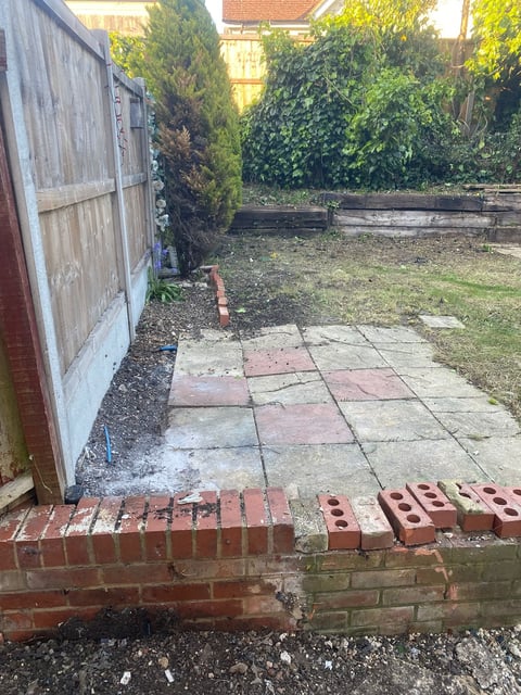 Overgrown garden patio with weathered brick edging, metal shed, and surrounding vegetation