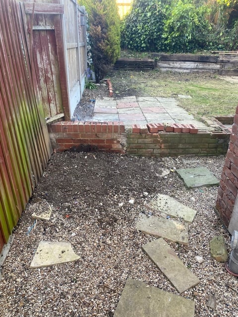 Overgrown backyard garden with gravel, scattered paving stones, brick walls, corrugated metal shed, and dense vegetation
